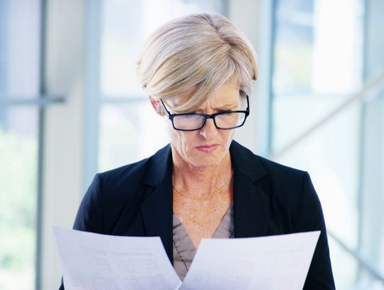 A businesswoman wearing glasses compares two printed documents while standing in a bright modern office.