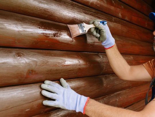 A man wearing gloves and a blue cap brushing paint onto a glossy brown log cabin wall while steadying a lower log.