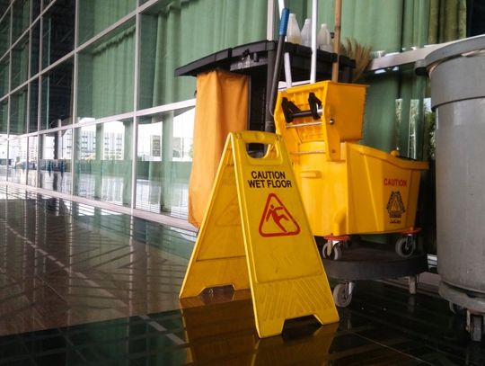 A business hallway with a cleaning supply cart and a wet floor sign. There is a yellow mop system and a trash can.