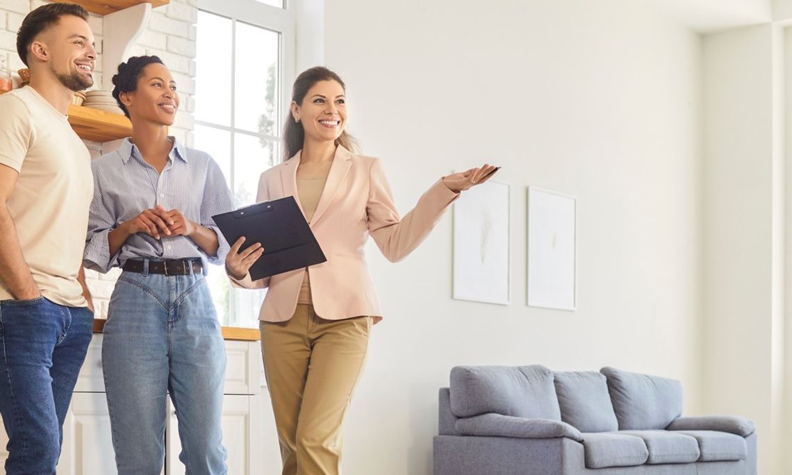 A female realtor holds a clipboard while showing the interior of a home to a young man and woman.