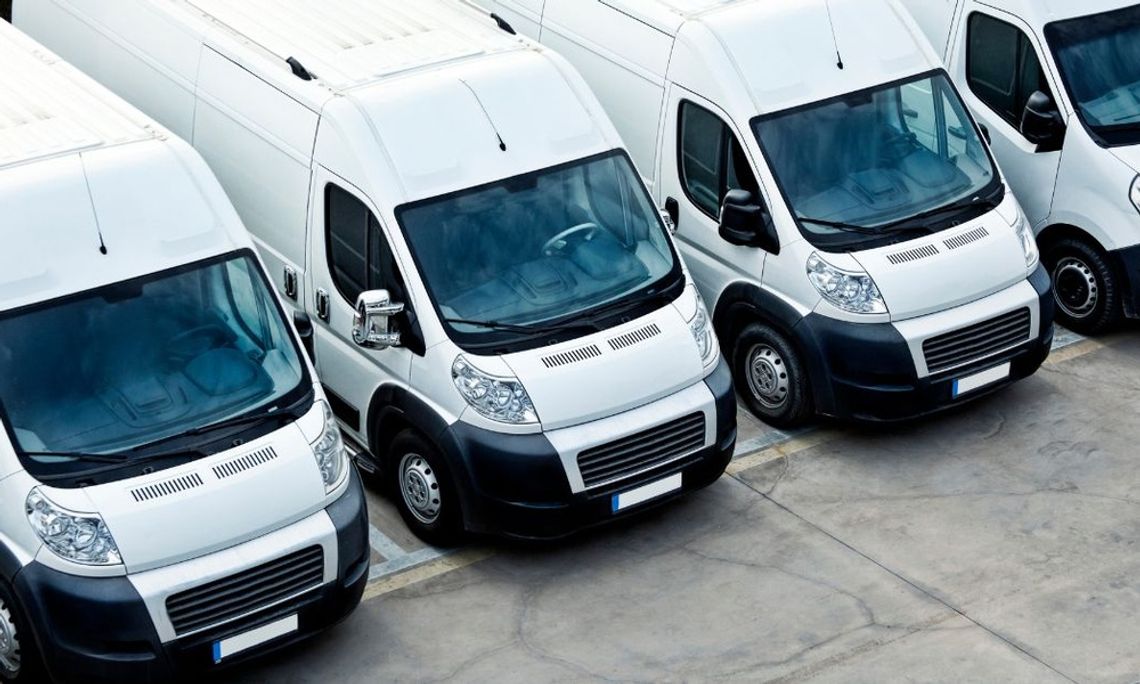 A fleet of four identical service vans sits in a paved parking lot. The white vans are parked in an orderly row.