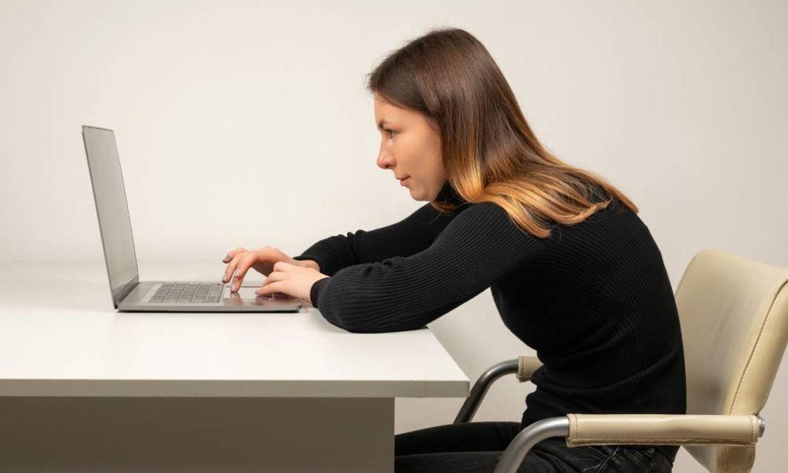A brown-haired woman in a black turtleneck and jeans slouched over a white desk while working on a laptop.