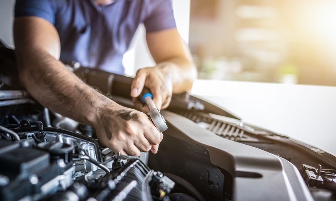 A close-up of a man in a T-shirt leaning over a car's engine and using a wrench on an exposed part.