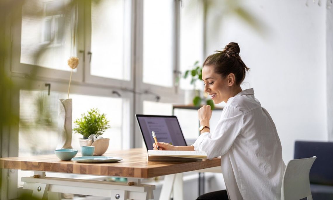 A woman in a white button-down shirt smiles as she writes in a notebook beside an open laptop on a wood desk.