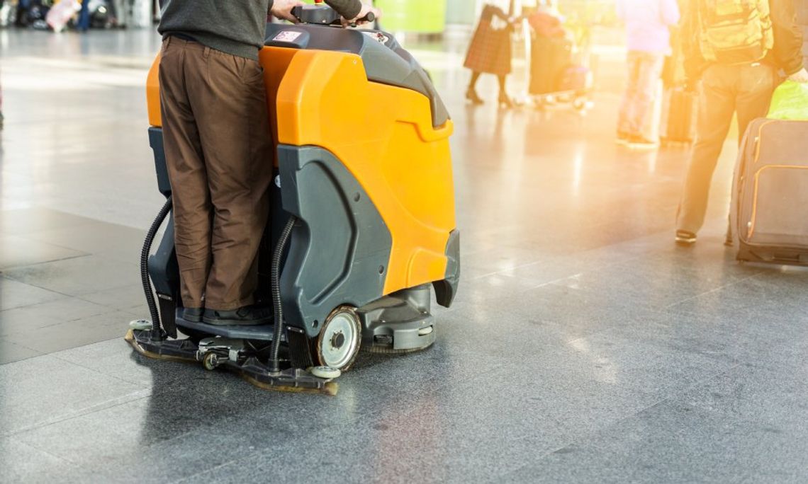 A custodian operating an industrial ride-on yellow floor cleaner across an expanse of tiled floor.