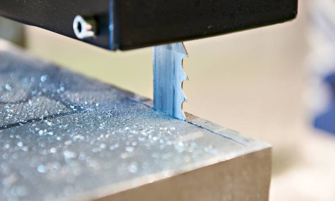 A close-up of a vertical band saw cutting a piece of metal. There are small metal shavings surrounding the blade.