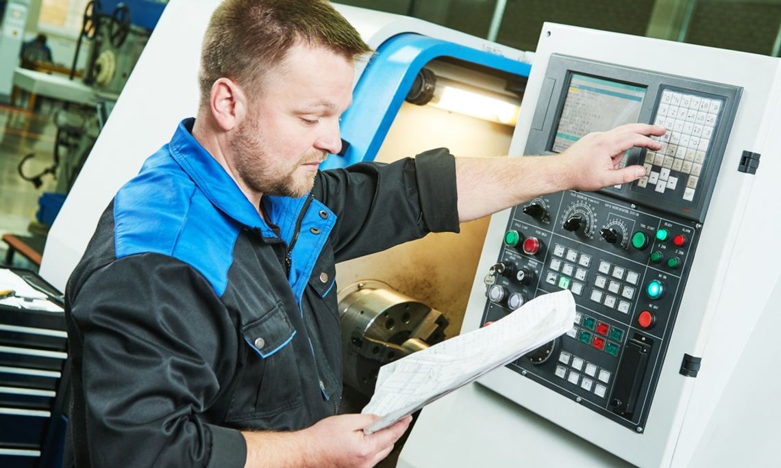 A machine operator is reading a manual while pressing machine buttons. He wears a black and blue shirt.