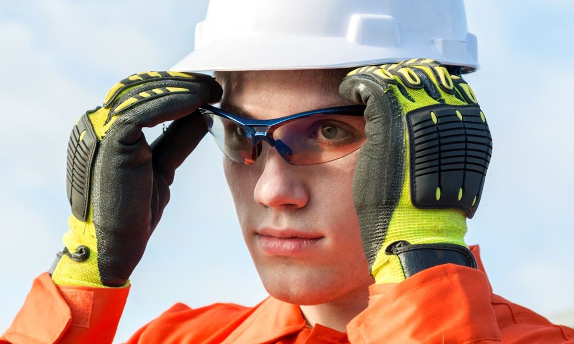 A young man in a white helmet and black and neon green work gloves is putting on blue safety goggles.
