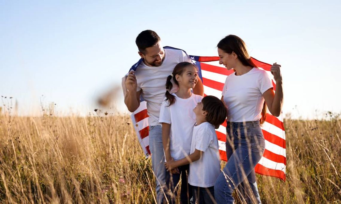 A family of four is standing outside on a sunny day. They are holding a large American flag over their shoulders.