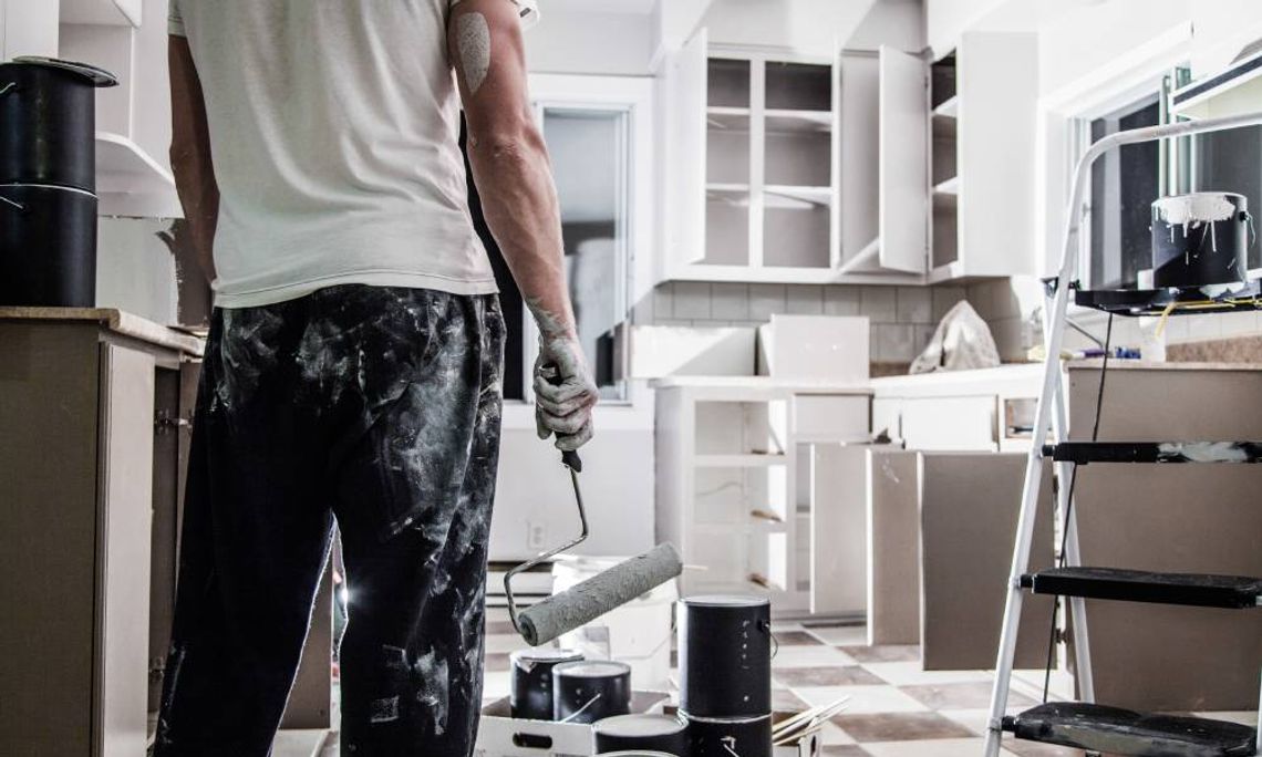 A male homeowner, covered in paint and holding a paint roller, standing in front of his kitchen in between renovations.