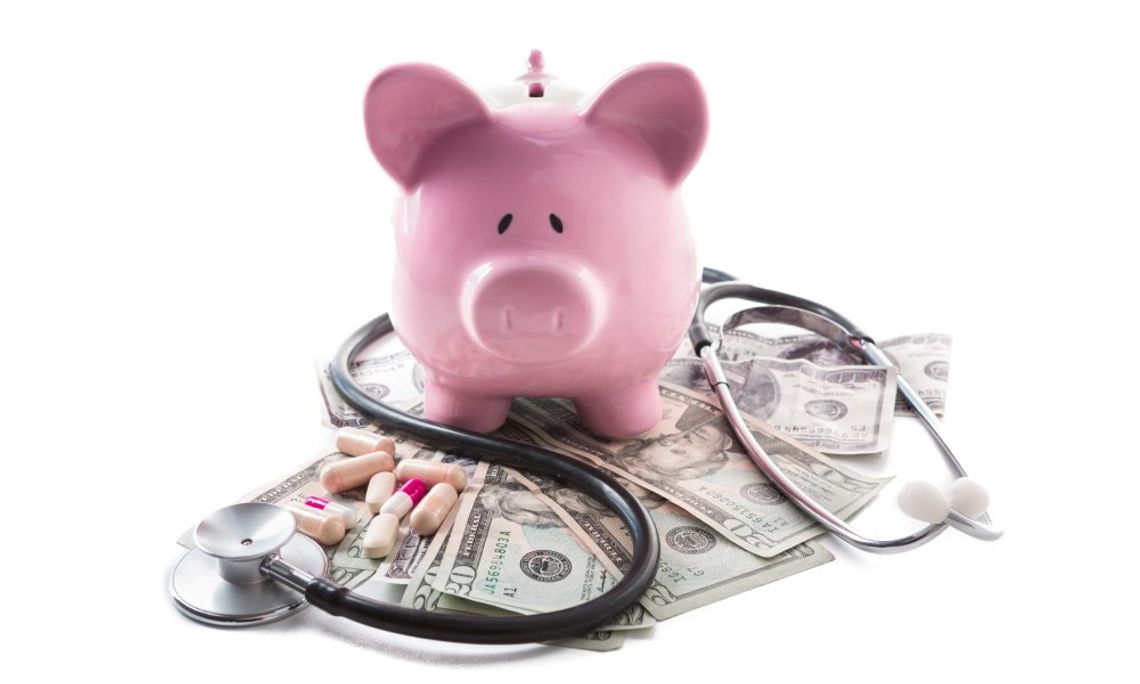 A pink piggy bank is sitting on top of a stack of money, a stethoscope, and a handful of medication.