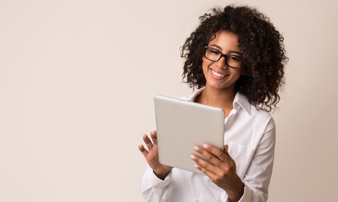 A woman with black curly hair standing against a beige background and smiling while looking at a silver tablet in her hands.