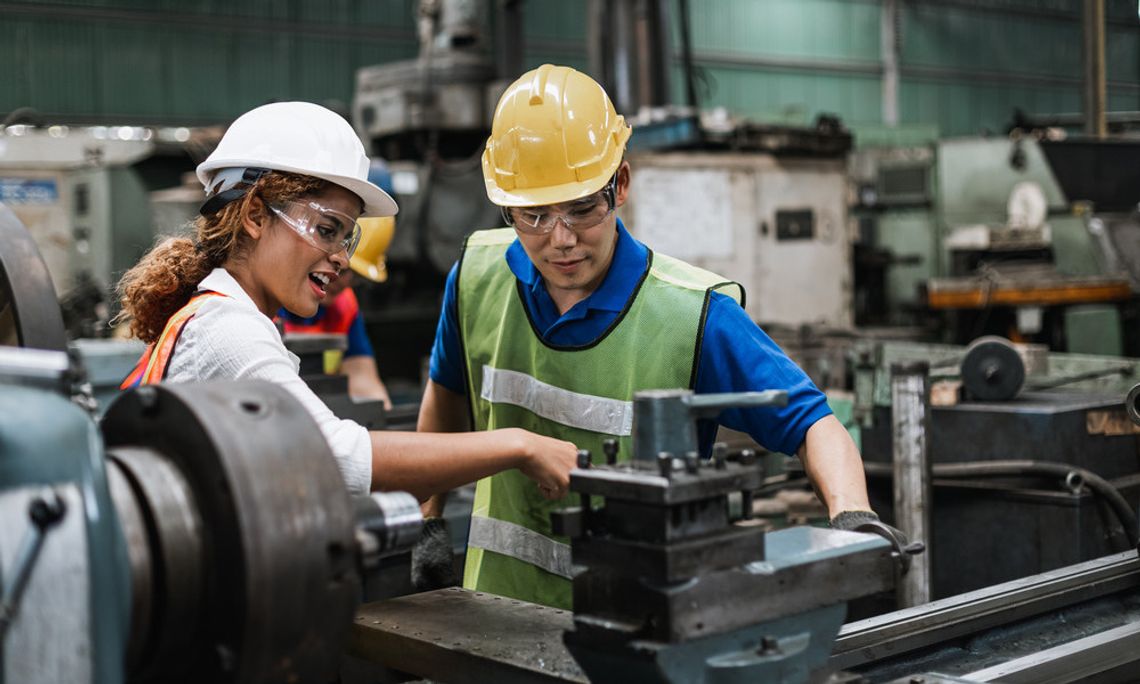 A man and a woman stand in front of a machine in a factory. Both wear hard hats, safety glasses, and safety vests.