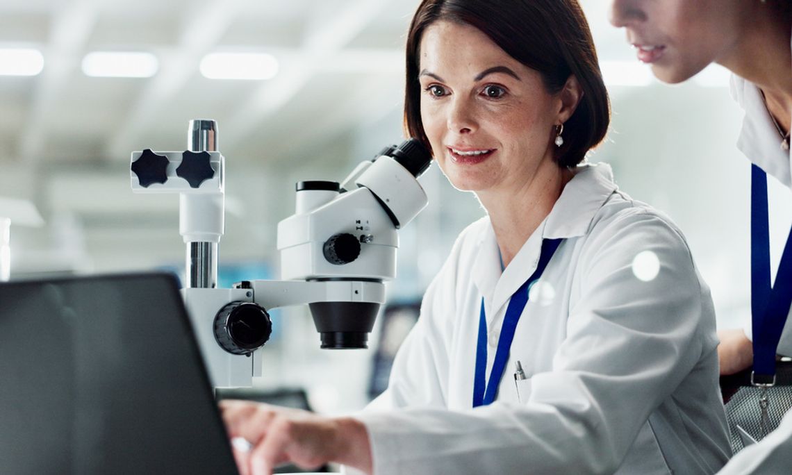 A scientist sits in a laboratory next to a microscope and points to a laptop screen while another scientist looks on.