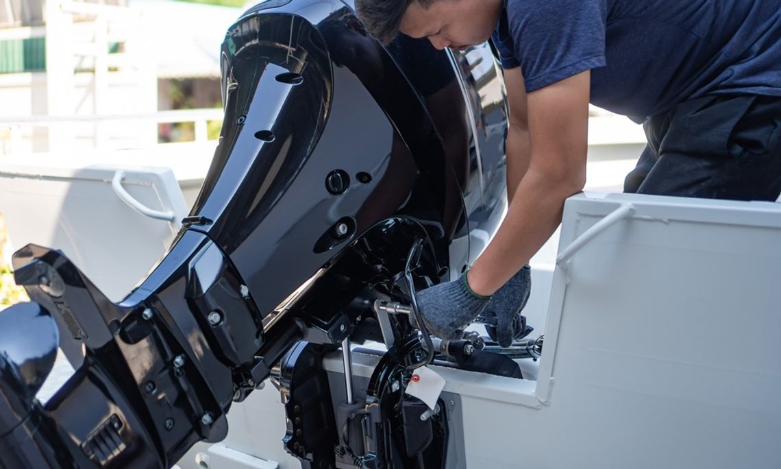 A man wearing gloves and a black t-shirt is bending over the back of the boat to work on the large, sleek black engine.