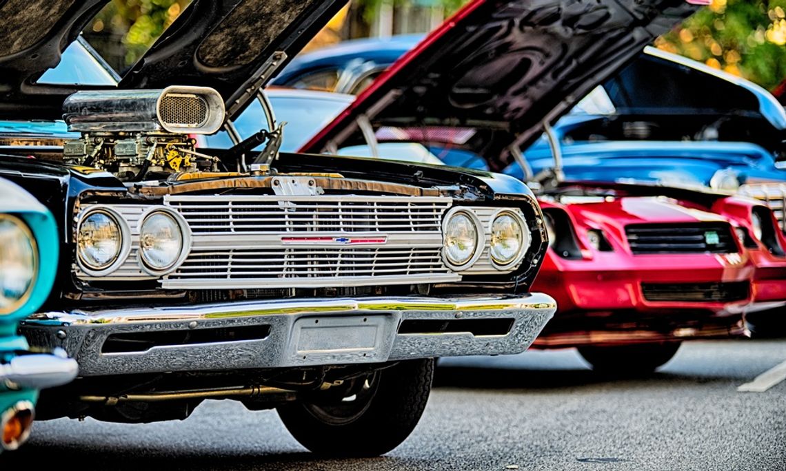 A lineup of multiple classic cars, all with bright, metallic finishes at a car show. Each car has its hood popped open.