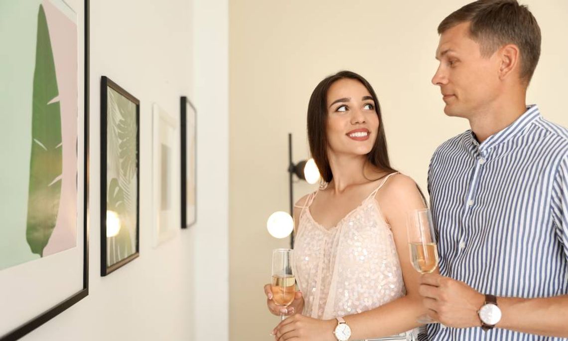 A young couple looking at each other and holding champagne flutes as they stand in front of a gallery exhibition.