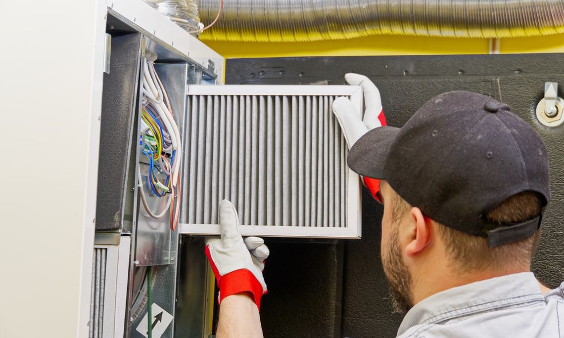 A man wearing a black hat and white and red gloves replacing the white filter in a ventilation system.