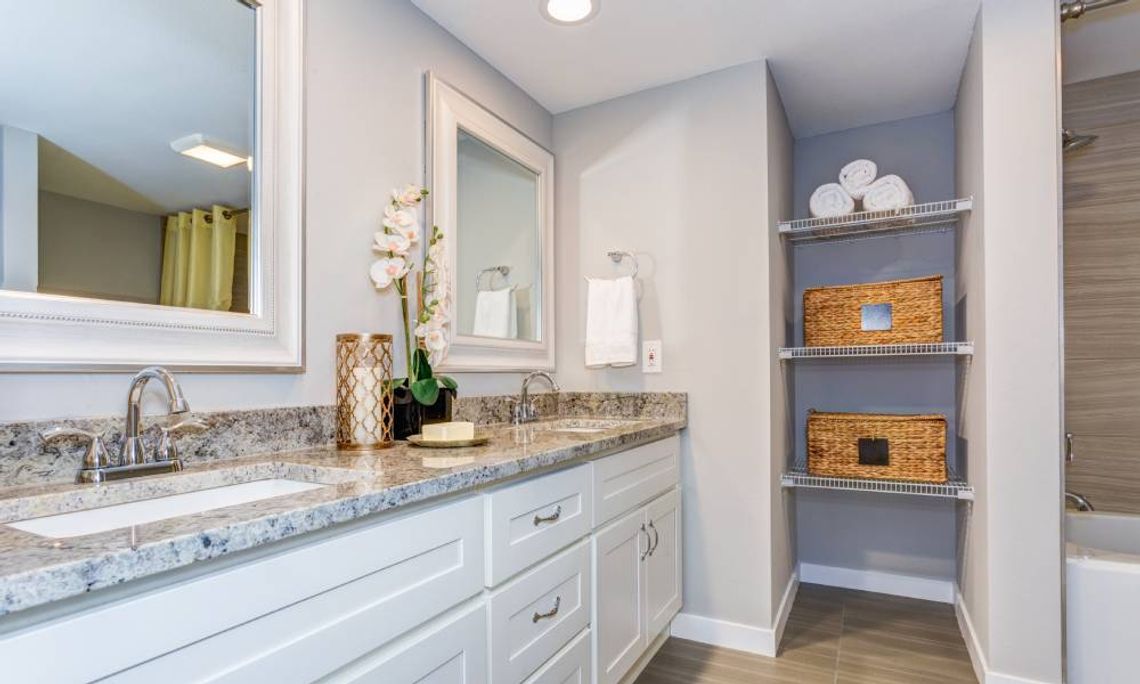 A home's bathroom with a granite countertop, two sinks, two mirrors, recessed shelving, and brown laminate flooring.