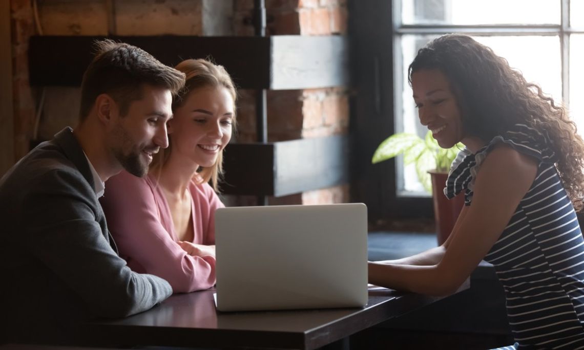 A woman sits with a couple at a table and shows them something on a laptop. What they are seeing makes them happy.