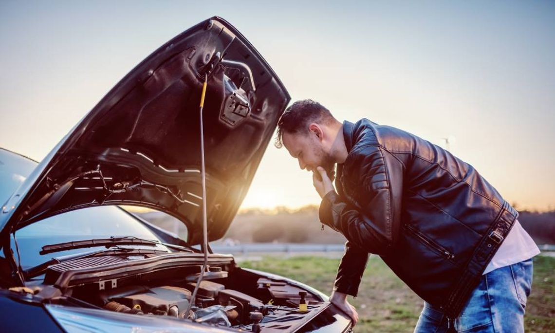 A young man in a leather jacket is looking at the engine of his car under the hood. There is a sunset in the background.