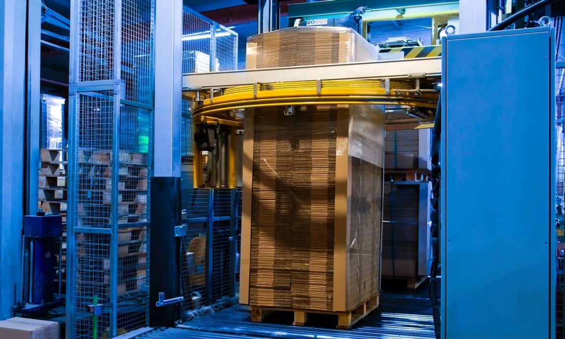 A blue stretch wrapping machine with a steel cage in the middle of wrapping a tall pallet of corrugated cardboard.