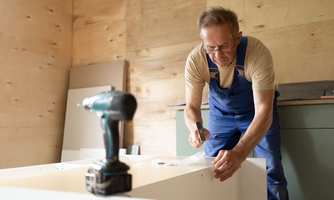 An older male in a tan shirt and denim overalls assembling a wooden piece of furniture in a workshop.