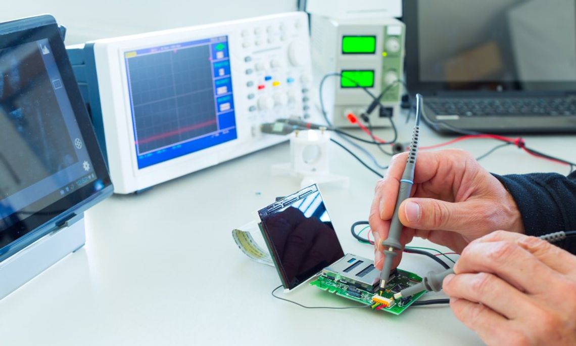 A technician's hands calibrate a piece of technology equipment on a table. A laptop lies on the surface nearby.