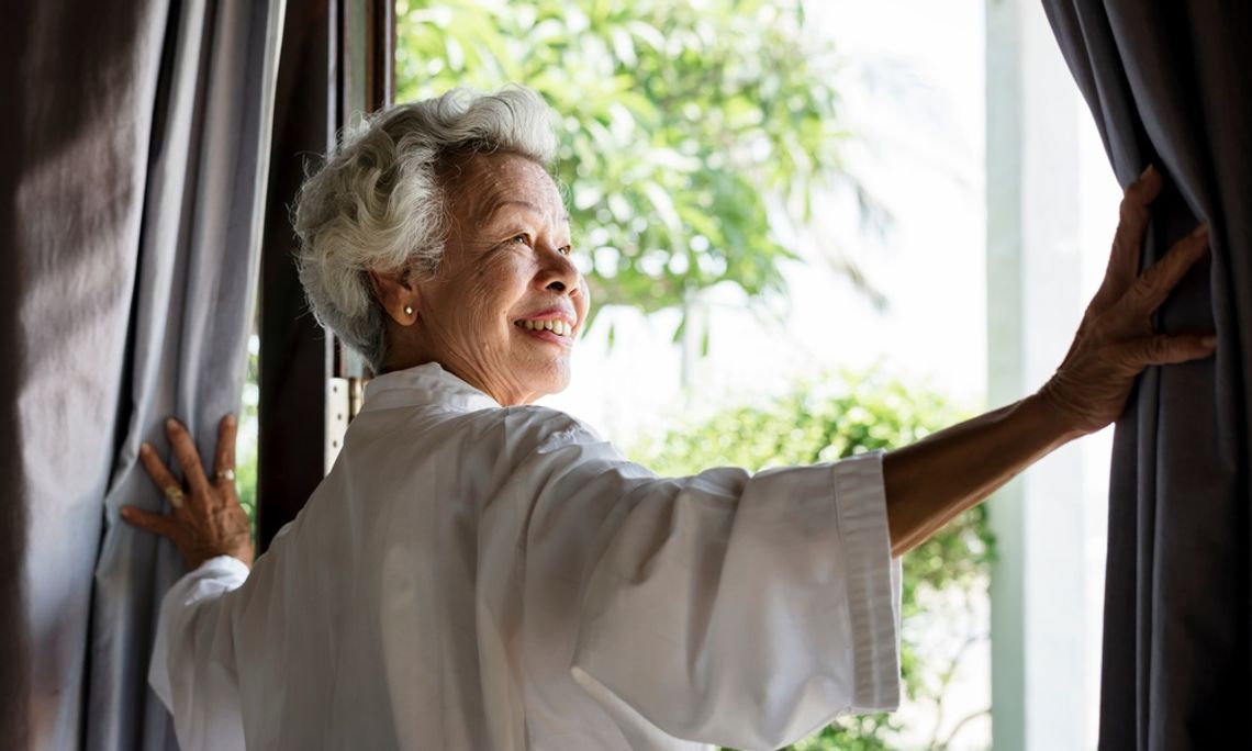 A woman smiles as she opens the curtains wide in her home, letting sunlight in. Trees are visible through the window.
