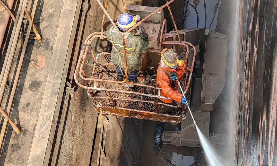 5 Types of Surfaces You Can Clean Using Hydroblasting Two people wearing protective gear are in a boom lift and using a hydroblasting machine to clean a surface.