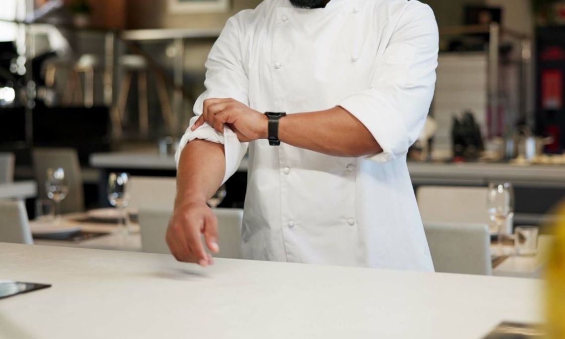 A man in a white chef’s jacket rolls up his sleeve while standing behind a white countertop in a dining area.