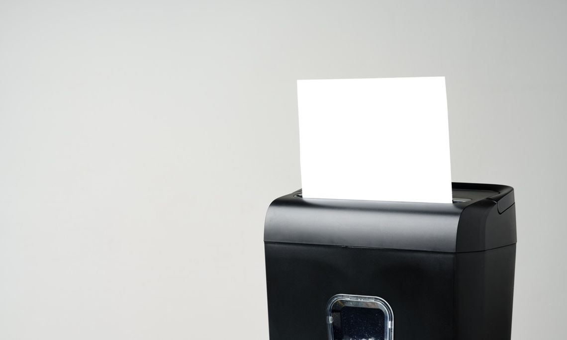 A blank piece of white paper sticks out of the top of a black shredder, which sits against a gray background.