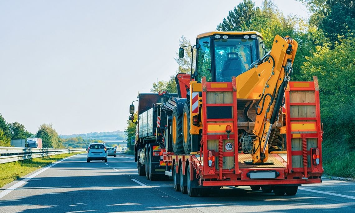 A large, heavy-duty truck is hauling heavy equipment and a tractor while driving on a busy highway.