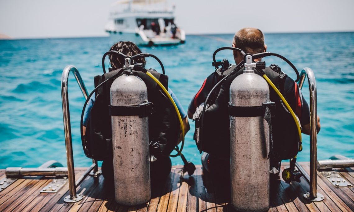 Two scuba divers sit on the edge of a wooden boat platform with their tanks and equipment, facing the blue ocean.