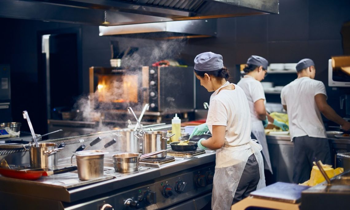 Several people in chef coats stand with their backs turned while working at counters in a commercial kitchen.