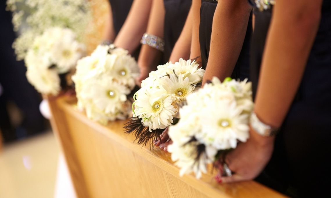 Bridesmaids in black dresses hold bouquets of white daisies with yellow centers while standing beside a wooden pew.