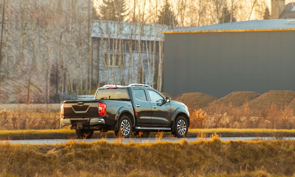 9 Battery Care Tips for Your High-Performance Truck A wide view of a black pickup truck driving on a wet road past dry grass, leafless trees, and industrial buildings.