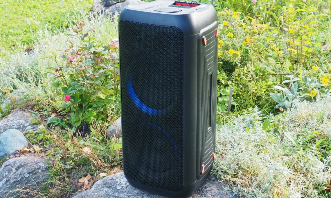A black rectangular speaker sitting on a rock with grass and colorful wildflowers in the background.