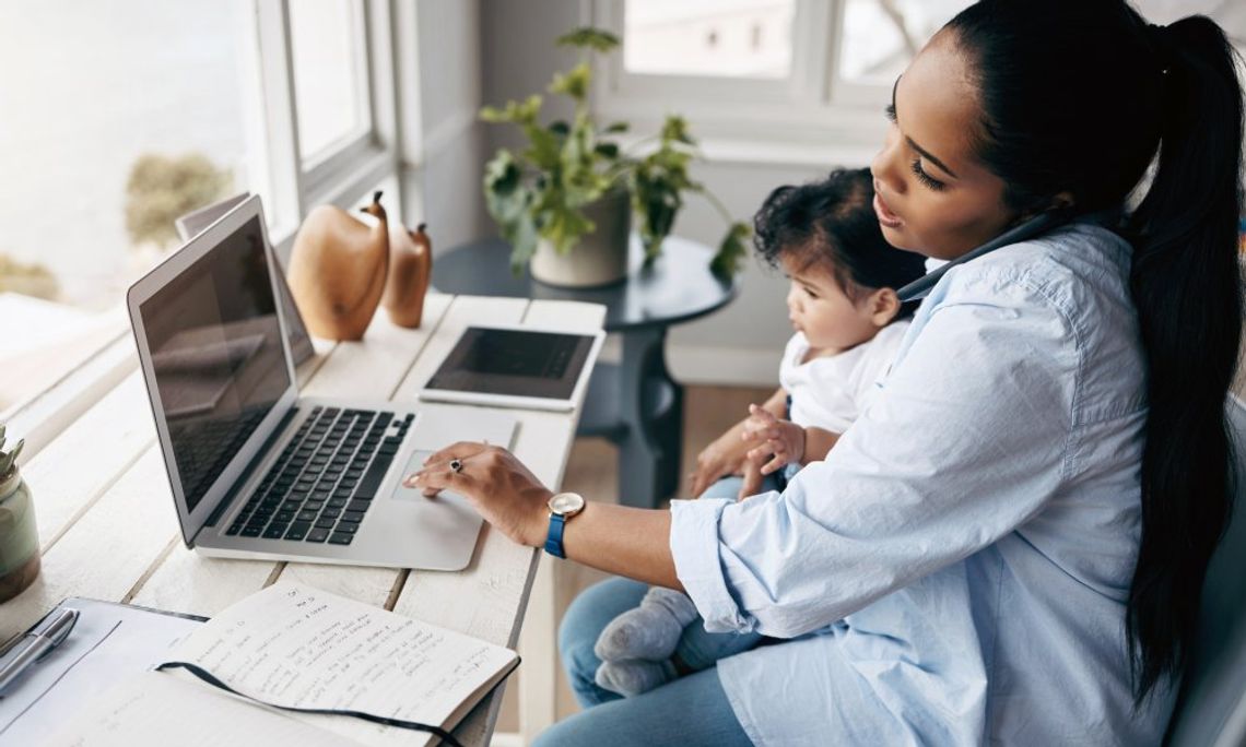 A mother holds her child in her lap as she talks on the phone and types on the computer.