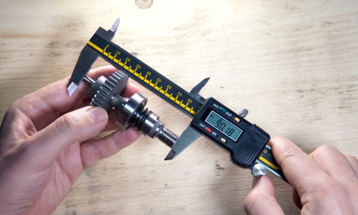 A machinist sitting at a wooden work table holding a digital caliper in both of their hands. The caliper reads 90.18.