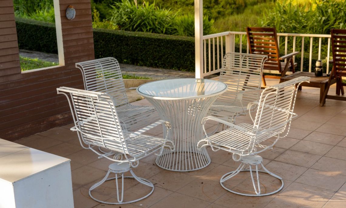 Benefits of Powder Coating for Patio Furniture A white metal patio table and four matching chairs on a covered porch, with greenery and wooden seating in the background.