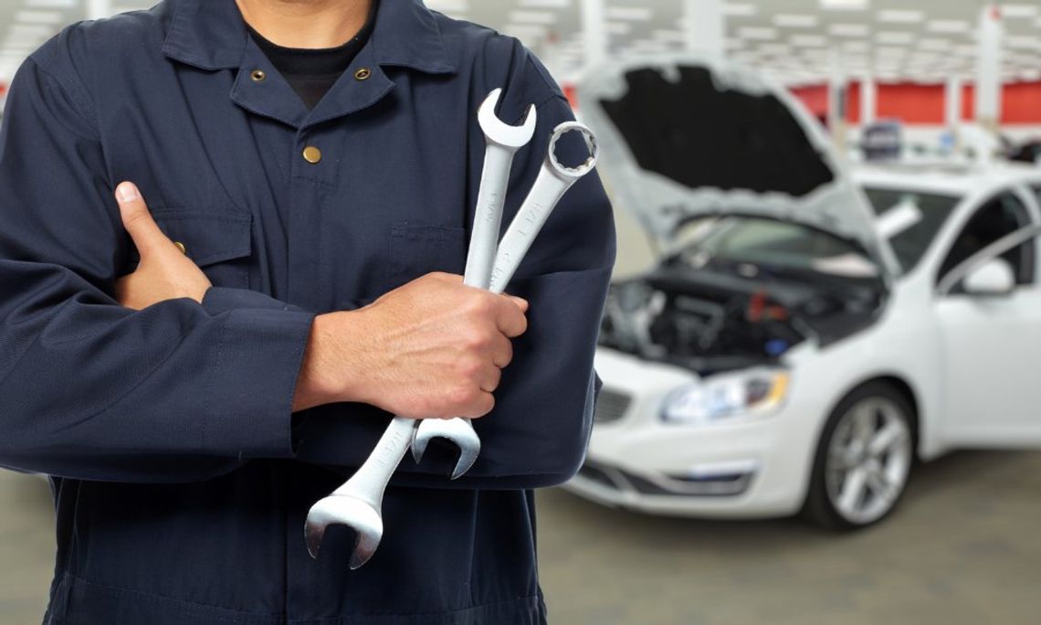 A person in a dark work uniform holding two wrenches in front of a car with an open hood inside a repair shop.