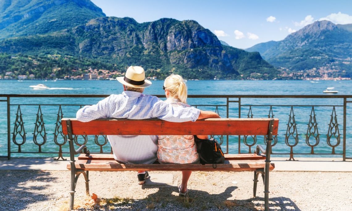 Best Romantic Getaway Styles for Couples Couple sits on a lakeside bench overlooking blue water and towering green mountains under a clear summer sky.