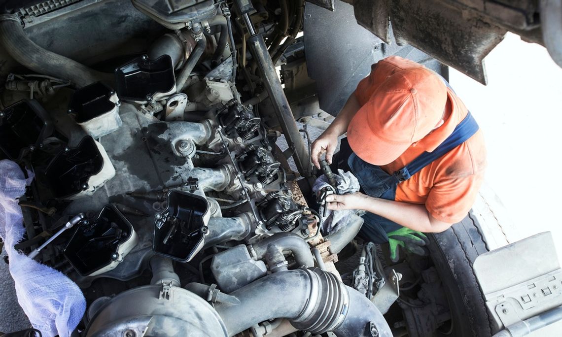 Overhead view of a mechanic in an orange hat, orange shirt, and overalls working on an open truck engine.