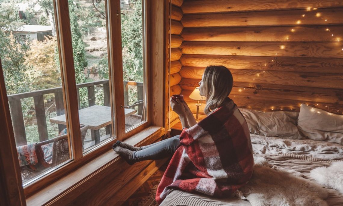 Best Ways To Unwind During a Cabin Vacation Woman wrapped in a red plaid blanket enjoys a warm drink beside large windows overlooking trees in a cozy log cabin.