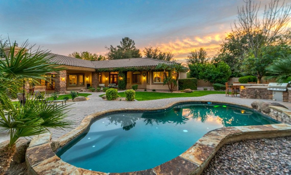 A luxurious backyard at sunset. There is a pool, an outdoor dining area, and pretty landscaping.