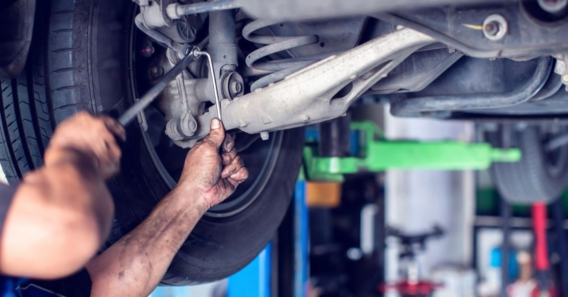 Car Maintenance: Repair vs. Rebuild vs. Replace A person's hands using wrenches on a raised vehicle's rear suspension near a tire inside an auto repair shop.