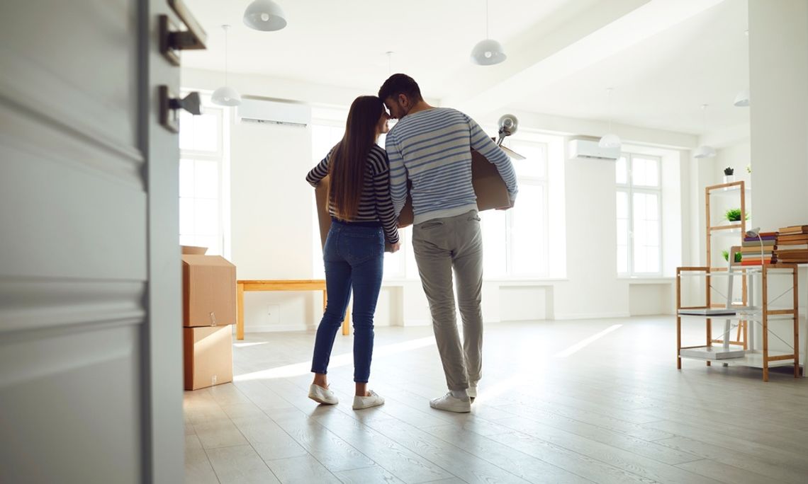 A man and a woman are standing in a room that is not furnished. They’re each holding cardboard boxes.