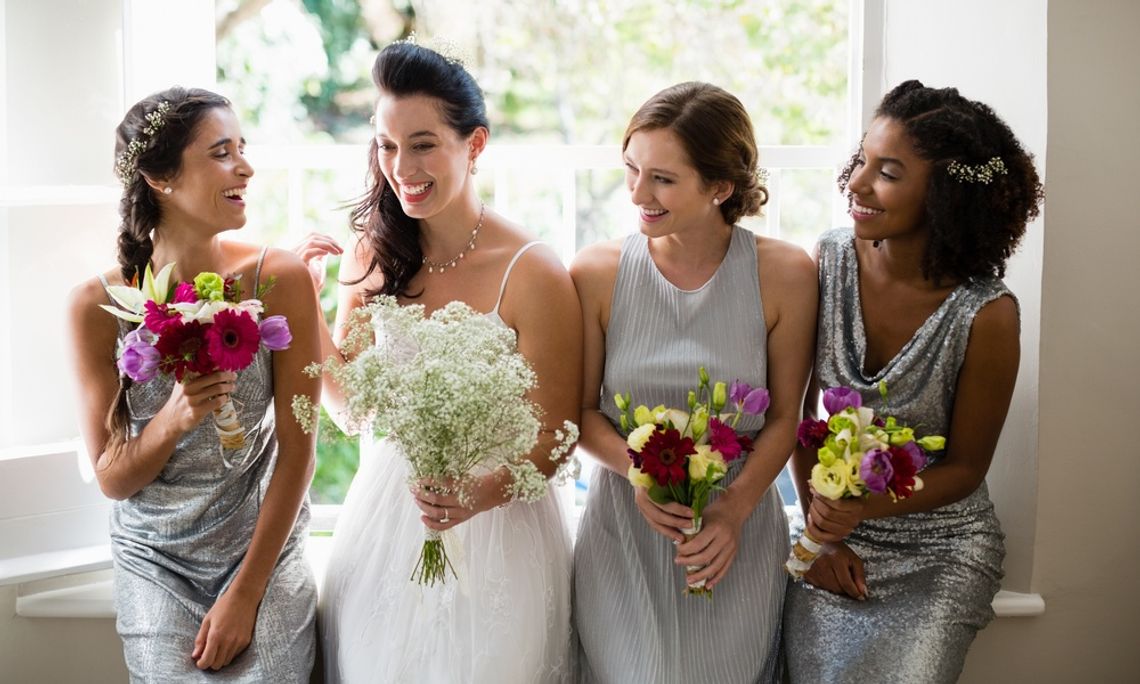 Choosing Bridesmaid Dresses for Real Life After the Wedding A woman in a white dress standing with three women in silver dresses holding bouquets near a bright window.