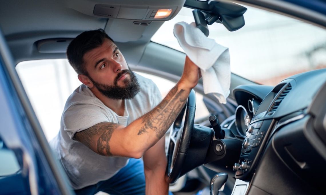 Cleaning Techniques: The Proper Way to Maintain Your Car A man with tattoos is kneeling inside of a car. He’s using a fabric cloth to clean the rear-view mirror.